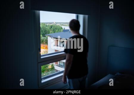 Mann und Fenster im dunklen Raum. Nachbar ausspionieren oder schnüffeln. Einsamkeit, Scham oder Melancholie. Deprimiert, traurig, launisch oder paranoid. Psychische Gesundheit. Stockfoto