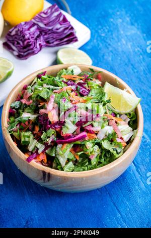 Frischer Gemüsesalat mit Purpurkohl, Weißkohl, Salat und Karotte in einer Holzschüssel auf blauem Holzhintergrund. Stockfoto