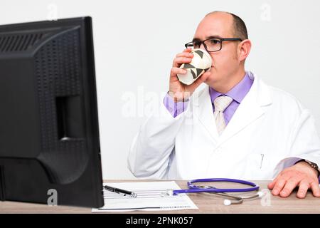 Ein kahlköpfiger, weißer Arzt mit Brille und weißem Mantel trinkt einen Kaffee zum Mitnehmen in seinem Büro vor einem Computermonitor. Stockfoto