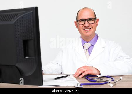 Ein kahlköpfiger Arzt mit Brille, der einen weißen Mantel trägt, der vor einem Tisch sitzt, wo ein Computermonitor, ein Dokument, ein Stift und ein Stethoscop sind Stockfoto