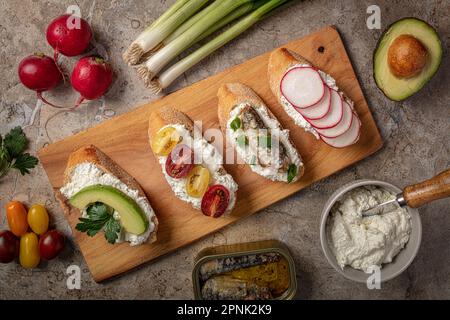 Frühstücksbrot mit Frischkäse und verschiedenen Gemüsesorten auf Holzbrett Stockfoto