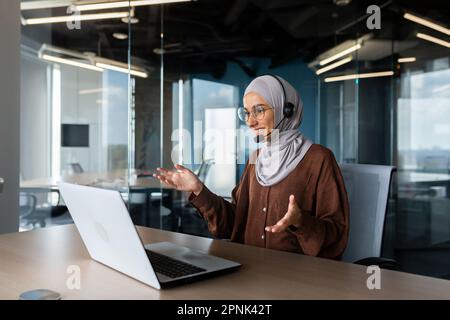 Online-Unterricht. Junge muslimische Frau in Hidschab und Headset, Lehrerin sitzt am Schreibtisch vor dem Laptop, erklärt, spricht, Gesten mit den Händen. Führt Fernunterricht durch. Stockfoto