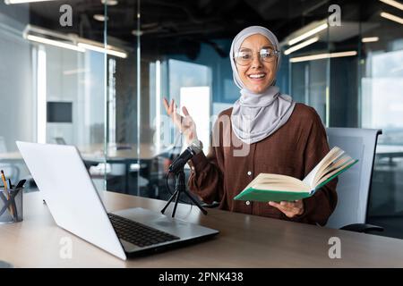 Online-Unterricht. Junge muslimische Frau in Hidschab-Lehrerin sitzt am Schreibtisch im Büro vor Laptop und Mikrofon und hält Buch in den Händen. Führt Fernunterricht durch. Stockfoto
