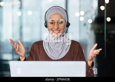 Nahaufnahme. Online-Unterricht. Porträt einer jungen arabischen Frau in Hijab und Headset, Lehrerin, die vor dem Laptop sitzt, erklärt, spricht, Gesten mit den Händen macht. Führt Fernunterricht durch. Stockfoto
