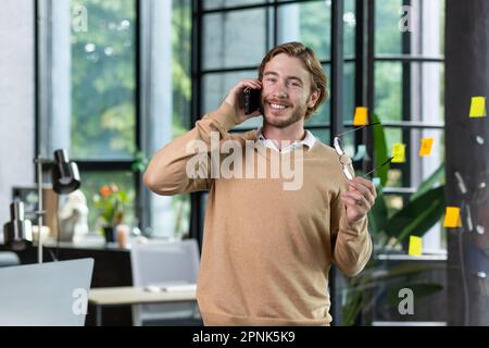 Ein junger Freiberufler steht im Büro und telefoniert. Er hält die Brille in der Hand, sieht in die Kamera und lächelt. Stockfoto