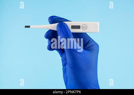 Die Hand eines Mannes in blauen Handschuhen trägt ein Thermometer auf blauem Hintergrund. Ein Arzt in blauen Latexhandschuhen zeigt ein elektronisches Thermometer. Stockfoto