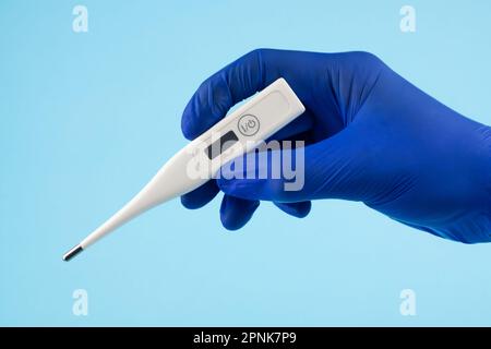 Die Hand eines Mannes in blauen Handschuhen trägt ein Thermometer auf blauem Hintergrund. Ein Arzt in blauen Latexhandschuhen zeigt ein elektronisches Thermometer. Stockfoto