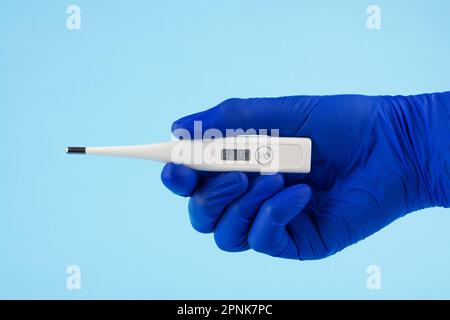 Die Hand eines Mannes in blauen Handschuhen trägt ein Thermometer auf blauem Hintergrund. Ein Arzt in blauen Latexhandschuhen zeigt ein elektronisches Thermometer. Stockfoto