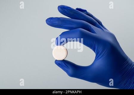 Männliche Hand in einem blauen medizinischen Handschuh mit einer weißen Pille auf grauem Hintergrund. Schließen Stockfoto
