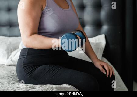 Mittellange Aufnahme einer Latina-Frau, die eine sitzende Übung auf ihrem Bett mit einer blauen Kurzhantel macht. Ich arbeite am Bizeps. Stockfoto
