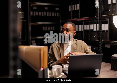 Ein afroamerikanischer Polizist, der mit Archivakten arbeitet und Informationen auf dem Laptop durchsucht. Ein Detective sitzt am Arbeitsplatz neben einem Regal mit Polizeiakten Stockfoto