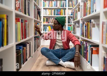 Ein nachdenklicher, afroamerikanischer Student, der in der Universitätsbibliothek Forschungslehrbücher auswählt. Stockfoto