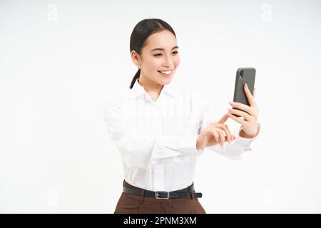 Eine schöne lächelnde Geschäftsfrau macht Selfie auf dem Handy, macht ein Foto auf dem Smartphone, nimmt Videos auf, steht auf weißem Hintergrund Stockfoto