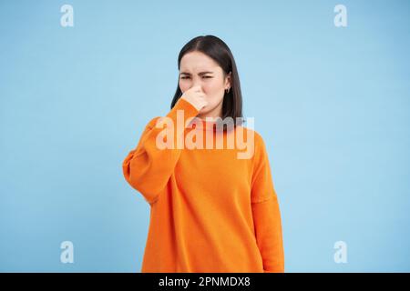 Irgendwas stinkt hier. Junge Asiatin schließt ihre Nase mit Fingern von schlechtem Geruch, trägt orangefarbenes Sweatshirt, steht über blauem Hintergrund Stockfoto