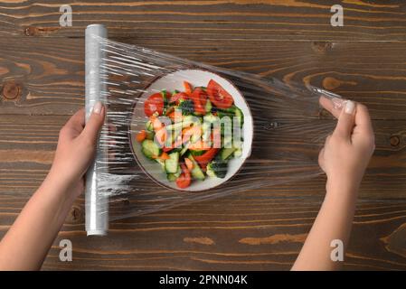 Weibliche Hände wickeln Schüssel mit Salat in Stretchfolie auf Holzhintergrund Stockfoto