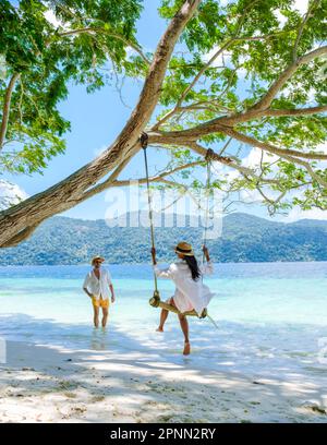 Ein paar Männer und Frauen sind am Strand mit einer Seilschaukel auf Koh Lipe Island Süd Thailand mit türkisfarbenem Meer und weißem Sandstrand bei Ko Lipe. Tarutao-Nationalpark, Insel Koh Rawi Stockfoto