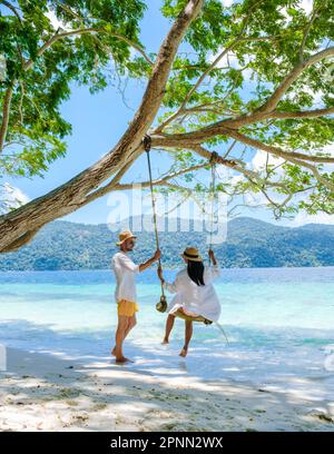 Ein paar Männer und Frauen sind am Strand mit einer Seilschaukel auf Koh Lipe Island Süd Thailand mit türkisfarbenem Meer und weißem Sandstrand bei Ko Lipe. Tarutao-Nationalpark, Insel Koh Rawi Stockfoto