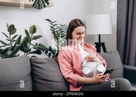 Attraktive, junge Schwangere, die Kopfhörer auf dem Bauch trägt und allein auf dem bequemen Sofa zu Hause sitzt. Glückliche werdende Mutter, die Musik in gemütlicher Livi hört Stockfoto