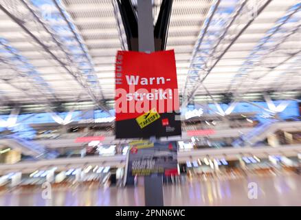 20. April 2023, Hamburg: Ein Poster aus Verdi mit dem Text „Warning Strike today“ hängt auf einer Anzeigetafel in Terminal 1 am Flughafen Hamburg. Die Gewerkschaft Verdi hat auf drei Flughäfen, darunter Hamburg, einen Warnstreik gefordert. Hintergrund für die Warnschläge sind die Verhandlungen zwischen Verdi und dem Bundesverband der Flugsicherheitsgesellschaften (BDLS) über Zeitzuschläge für Nacht-, Samstag-, Sonntag- und Urlaubsarbeit sowie eine bessere Tarifvereinbarung über die Überstundenvergütung für Sicherheits- und Servicepersonal auf kommerziellen Flughäfen. Foto: Christian Charisius/dpa Stockfoto