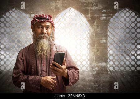 Ein muslimischer Mann mit Bart, der Keffiyeh mit Agial trägt und ein Handy mit blauem Himmelshintergrund benutzt Stockfoto