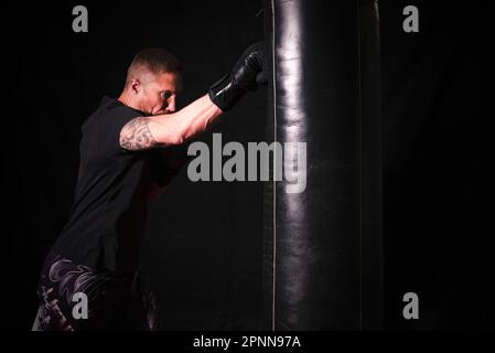 Ein weißer Mann, der ein schwarzes T-Shirt trägt und in einer Turnhalle eine hängende Boxtasche schlägt Stockfoto