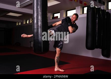 Ein weißer Mann, der ein schwarzes T-Shirt und Shorts trägt und eine hängende Boxtasche im Fitnessstudio schlägt Stockfoto