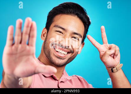 Glücklicher asiatischer Mann, Porträt und Friedenszeichen für Selfie, Profilbild oder soziale Medien vor blauem Studiohintergrund. Männlicher Einflussnehmer oder Vlogger Stockfoto