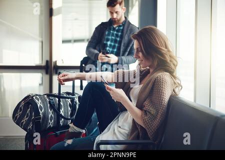 Ich verabschiede mich nur von der Familie. Ein junges Paar, das mit seinem Gepäck am Flughafen sitzt und dabei sein Handy benutzt. Stockfoto