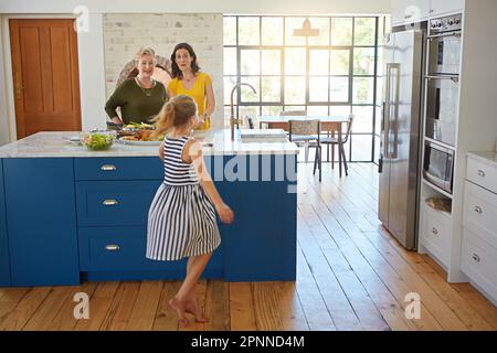 Sie freut sich auf eine gesunde Mahlzeit. Ein kleines Mädchen tanzt, während ihre Mutter und Großmutter im Hintergrund kochen. Stockfoto