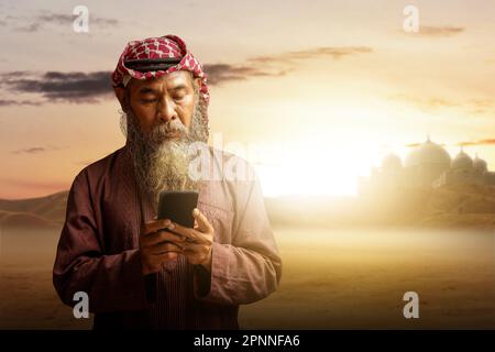 Ein muslimischer Mann mit Bart, der Keffiyeh mit Agial trägt und ein Mobiltelefon mit dem Hintergrund der Sonnenuntergangsszene benutzt Stockfoto