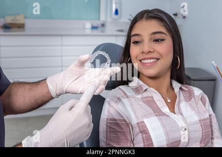 Entzückte hispanische Teenagerin mit langen dunklen Haaren in lässiger Kleidung, die lächelt, während sie auf transparente, vakuumgeformte Halterung in den Händen des Ernteguts blickte Stockfoto