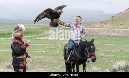 Issyk Kul, Kirgisistan - Mai 2022: Adlertrainer hilft einem Touristen, seinen goldenen Adler zu halten. Traditioneller Tourismus in Kirgisistan Stockfoto