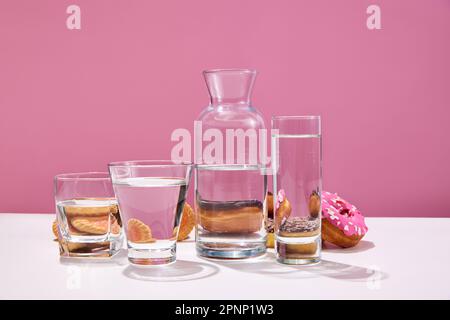 Stillleben mit Backkeksen, Donuts mit Zuckerglasur und Gläsern mit Wasser auf einem weißen Tisch mit pinkfarbenem Studiohintergrund. Leckere Süßigkeiten Stockfoto