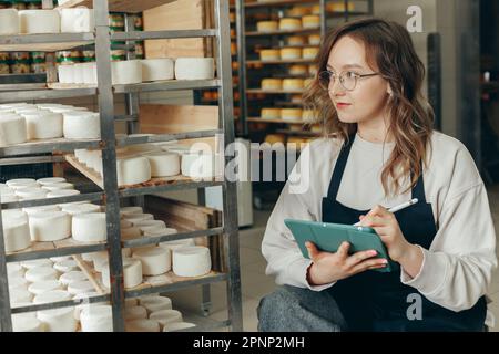 Junge Farm weibliche Besitzerin kontrolliert den Reifungsprozess von Ziegenkäseköpfen, die in den Regalen des Lagerhauses der Käserei platziert werden. Notizen mit Digital T schreiben Stockfoto