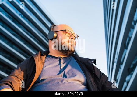 Ein Mann in Übergröße mit Brille, der Musik über Kopfhörer hört und das Telefon benutzt. Er sitzt im Freien mit modernem Bürogebäude im Hintergrund und in der Sonne Stockfoto