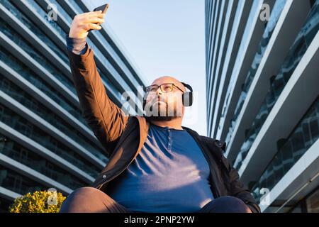 Ein Mann in Übergröße mit Bart und Brille macht Selfie, während er Musik über Kopfhörer hört. Er sitzt draußen mit einem modernen Bürogebäude auf dem Rücken Stockfoto