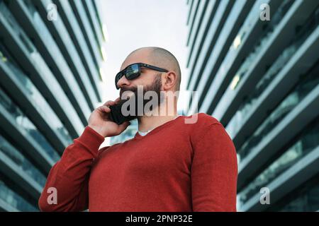 Ein bärtiger Mann in einer Sonnenbrille, der sich draußen in den modernen Bürogebäuden mit seinem Handy unterhält. IT-Mitarbeiter oder Freiberufler, die Mobiltelefone verwenden. Schließen Stockfoto
