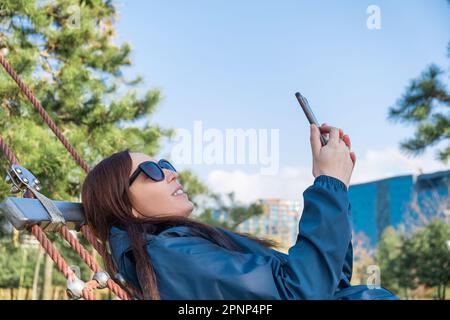 Wunderschöne junge Frau, die mit ihrem Smartphone im Internet surft, soziale Medien nutzt und lächelt, liegt auf einer Hängematte im Park. Moderner Lifestyle, Verbindung, s Stockfoto