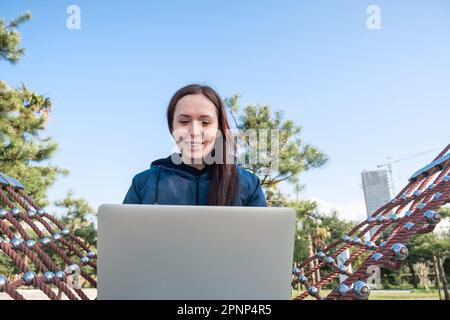 Junge weiße Frau, digitaler Nomade, Freiberufler, der im Freien arbeitet, sitzt in einer Hängematte, benutzt einen Laptop, lächelt. Fröhlicher weiblicher Chat auf Zoom oder Stockfoto