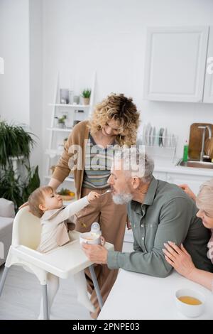 Ein kleines Mädchen mit Löffel füttert einen bärtigen Großvater in der Nähe einer lächelnden Familie in der Küche, Bild von einem Stock Stockfoto