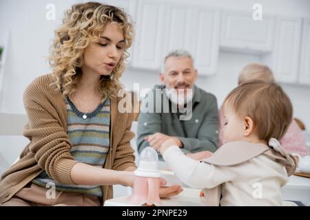 Frau füttert kleine Tochter mit Frühstück in der Nähe reifer Eltern, die auf einem verschwommenen Hintergrund in der Küche lächeln, Stockbild Stockfoto