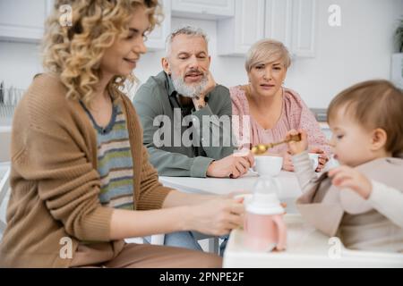 Ein Paar mittleren Alters, das sich die glückliche Tochter anschaut, die ein kleines Mädchen mit Frühstück füttert, in der Küche, Stockbild Stockfoto