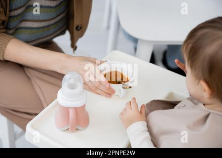 Kurze Aussicht auf eine Frau, die eine Schüssel mit Püree in der Nähe einer Kleinkindertochter hält, die während des Frühstücks auf einem Babystuhl sitzt, Stockbild Stockfoto