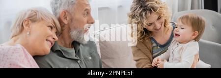 Verblüffter bärtiger Mann, der die Enkeltochter eines Kleinkindes in der Nähe der Familie auf der Couch im Wohnzimmer ansieht, Banner, Stockbild Stockfoto