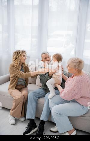 Ein bärtiger Mann hält eine Enkeltochter in der Nähe von Frau und glücklicher Tochter auf dem Sofa im Wohnzimmer, Bild der Aktie Stockfoto