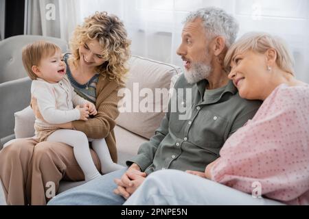 Eine lächelnde Frau, die mit einer kleinen Tochter in der Nähe glücklicher Eltern auf der Couch im Wohnzimmer sitzt, Bild der Aktie Stockfoto