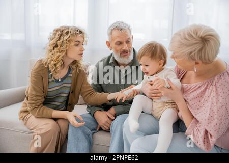 Lockige Frau beruhigt Baby in der Nähe von erwachsenen Eltern auf dem Sofa im Wohnzimmer, Stockbild Stockfoto