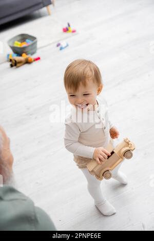 Blick aus einem großen Winkel auf ein überglücklich gewordenes Kind, das mit einem Spielzeugauto in der Nähe eines verschwommenen Großvaters im Wohnzimmer steht, Stockbild Stockfoto