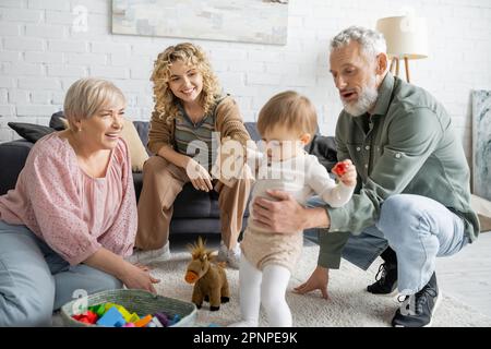 Lächelnde Frauen, die einen bärtigen Mann anschauen, der mit seiner Enkelin auf dem Boden im Wohnzimmer spielt, Stockbild Stockfoto