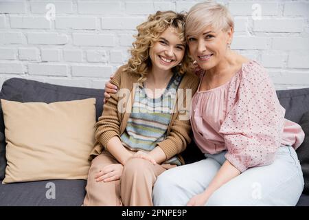 Glückliche Erwachsene Frau mit fröhlicher blonder Tochter, die auf der Couch im Wohnzimmer sitzt und auf die Kamera schaut, Stockbild Stockfoto